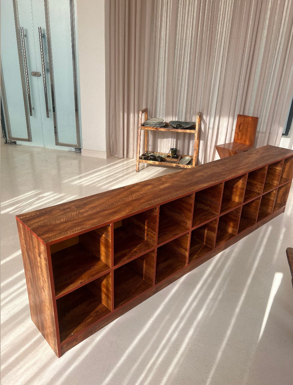 A long, low wooden storage shelf with multiple cubby compartments, shown in a modern interior room with sunlight and  shadows.
